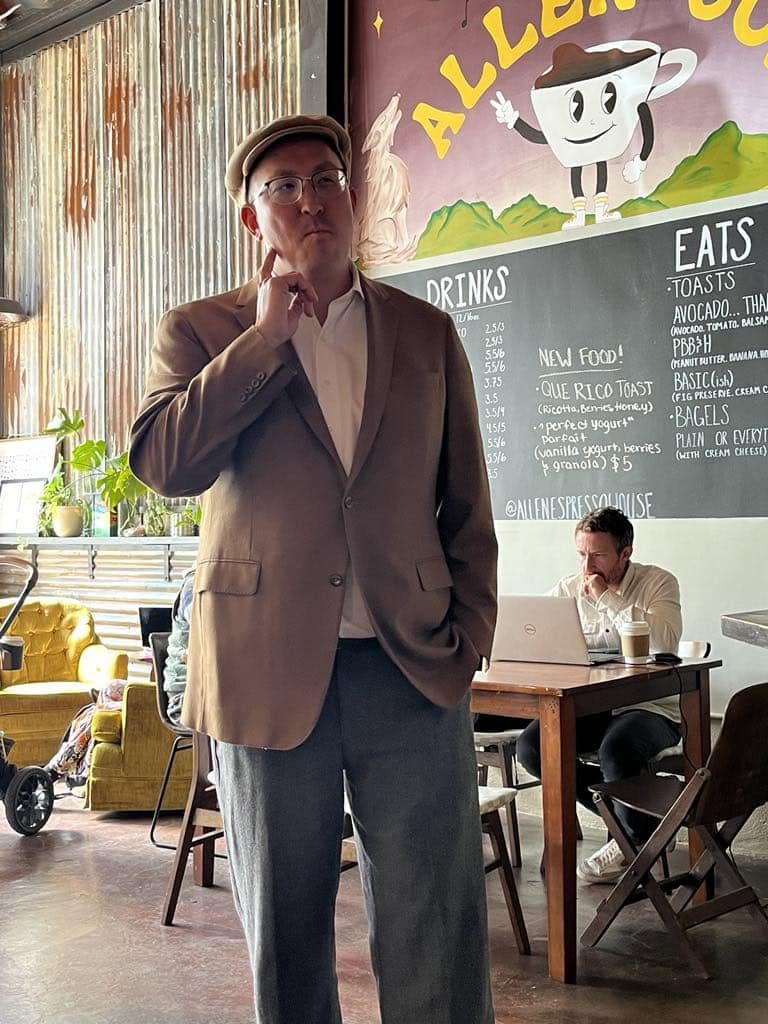 Nick Mueller standing in a café wearing a cap, blazer, and glasses